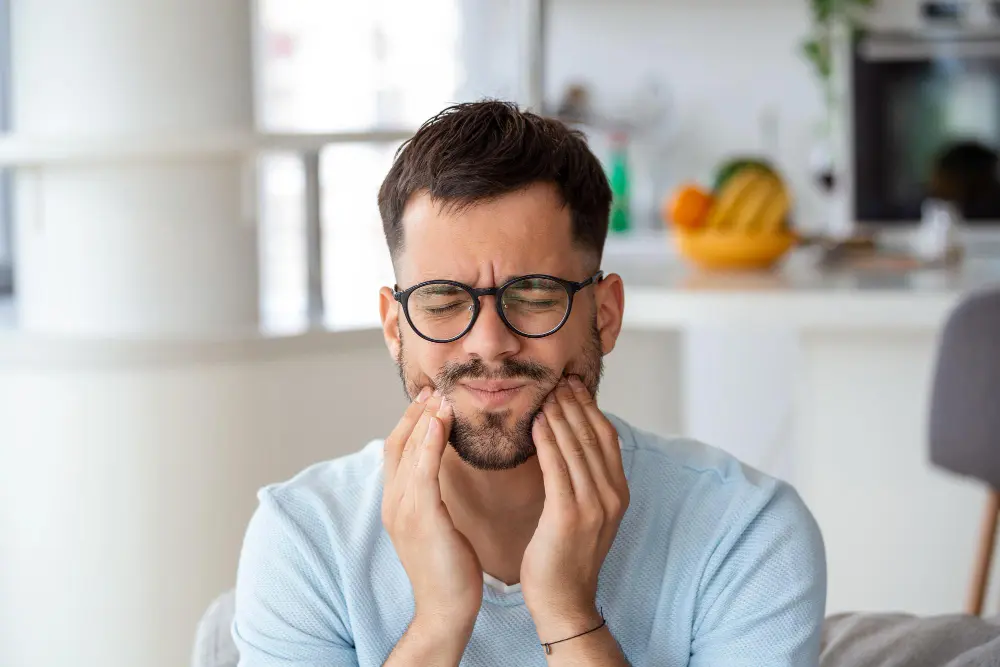 Young man holding his jaw in pain while sitting on the couch.